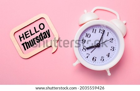 On a delicate pink background, a white alarm clock and a wooden frame with the text HELLO THURSDAY Royalty-Free Stock Photo #2035559426