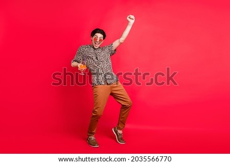 Full size photo of young excited cheerful man raise fist in triumph dancing drink alcohol cocktail isolated on red color background