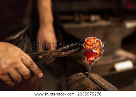Making iron, metal flower. Bearded man, blacksmith manually forging the molten metal on the anvil in smithy with spark fireworks. Concept of labor, retro vintage occupation, family business Royalty-Free Stock Photo #2035575278