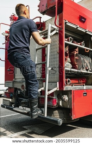 Dangerous work of firefighter. Male firefighter dressed in uniform climbing ladder in firefighting truck with specific equipment at fire station. Concept of job, career, safety, resque works.