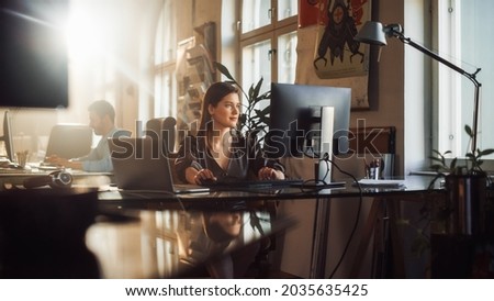 Two Stylish Employees Working on Computers in Creative Agency in Loft Office. Beautiful Manager Typing Correspondence Emails. Sunny Renovated Space with Plants, Artistic Posters and Big Windows. Royalty-Free Stock Photo #2035635425
