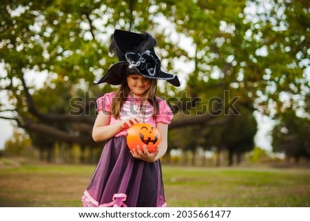 Portrait of little girl in witch Halloween costume and black hat tasting candy outdoor in forest with candy bucket