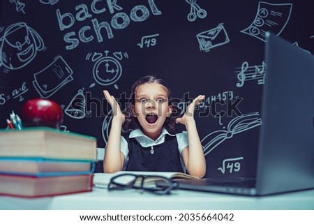 A sweet girl schoolgirl sits at the table with laptop, books and a chalkboard with school formulas on background. Works homework amazed to put her hands on head, opening mouth wide. 