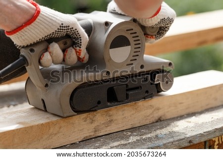 Carpenter sanding wood with belt sander in the garden. Woodworking carpentry. Close-up. Man in the garden sanding wooden planks. DIY home improvement, restoration, carpentry concept