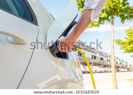 Woman plugging in the charger into a socket of her white electric car at a charging station in the street Royalty-Free Stock Photo #2035683020