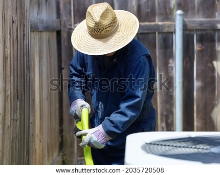 View of a man with a hat and coveralls doing yard work. Gardener using a weed eater.