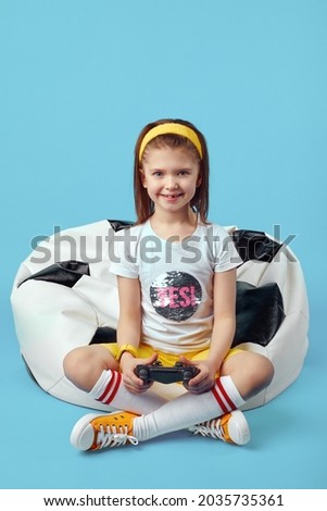 Full length photo of happy little girl sitting on bean bag chair, holding joystick and playing video game, wearing sport outfit, isolated over blue