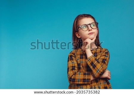 Cute caucasian schoolgirl in nerdy glasses and school uniform touching cheek and looking up while thinking against blue background
