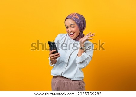 Excited Asian woman looking at smartphone getting good news over yellow background