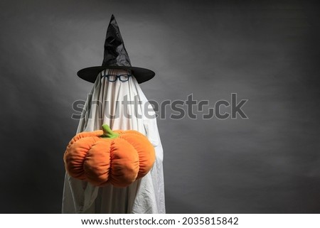 Costumed person with white sheet, glasses and witch hat carrying pumpkin. Halloween concept with space for text.