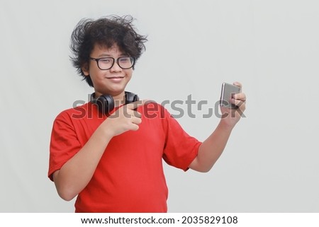 Portrait of Asian young boy in red t-shirt with glasses pointing on his smartphone. Isolated image on gray background