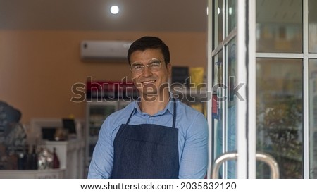 Young male owner expresses his warm welcome to customers for his local startup bakery or coffee shop in front of the shop. Smiling attractive owner with glasses stands for welcoming.