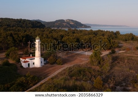 Aerial landscape before sunset featuring a brightly lit lighthouse against a background of dark trees on a Mediterranean seashore