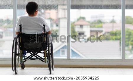 Back view of young Asian handicapped teenage boy without legs sitting on wheel chair and looking to outside of home with copy space on right. Royalty-Free Stock Photo #2035934225