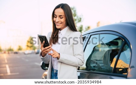 Successful smiling attractive woman in formal smart wear is using her smart phone while standing near modern car outdoors Royalty-Free Stock Photo #2035934984