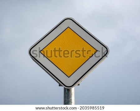 Metal road sign (Rectangular white with yellow square) "Main road" on metal pole against blue and white sky background