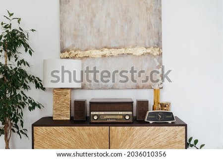 Home interior with vintage decor, living room design. Closeup of retro furniture with wooden decoration, drawer near white wall with poster. Old classic radio, clock, lamp on cabinet.