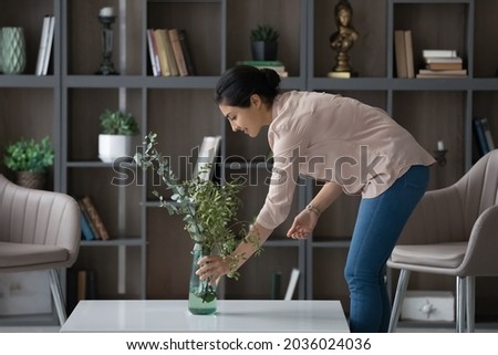 Smiling Indian woman taking care about green plant in cozy living room, pleasant attractive young female tenant renter decorating home, first own apartment, rent or mortgage, interior design concept Royalty-Free Stock Photo #2036024036