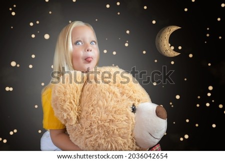 A little girl makes faces, hugging a teddy bear against the backdrop of a fabulous starry sky and the moon.