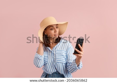 Beautiful thoughtful woman holds mobile phone in hand, doing online shopping, chatting, using application, wears straw hat, striped shirt, jeans with belt, on pink background. Summer emotions concept