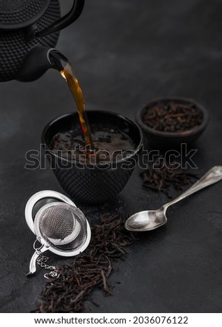 Pouring black tea from teapot to cup with strainer infuser and loose tea on black with silver spoon. Top view