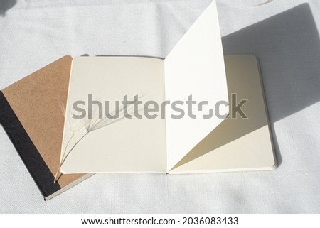 Modern summer stationery still life.Empty diary, notepad mockup with dandelion on white linen tablecloth. Long shadows of sunset in the sunlight. Top view