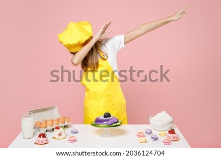 Teen girl chef cook confectioner baker wear yellow apron cap at table do dab hip hop dance hands move gesture youth sign isolated on pastel pink background Mousse cake food process workshop concept