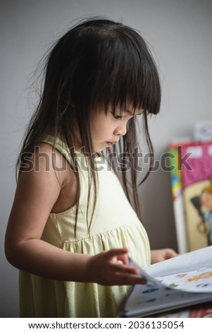 Selective focus is a 3-year-old Asian girl with long black hair sitting on a chair. She was paying attention to the picture book in his hand.