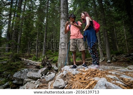 Interracial couple tourists smiling taking photo holding action camera with bag pack. Mobile smartphone selfie picture during adventure holiday hike trip in the forest. Summer lifestyle.