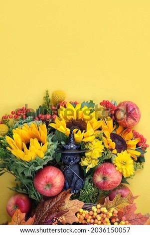 black cat figurine with witch hat, apples and autumn flowers on yellow background. fall season. symbol of autumn season, thanksgiving, halloween, mabon. top view. copy space