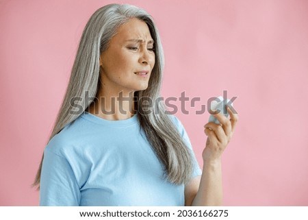 Doubting hoary haired Asian woman looks at sonic facial cleansing brush on pink background in studio. Mature beauty lifestyle