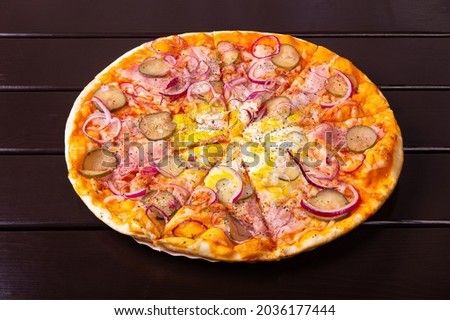 A whole rustic pizza with pickled cucumber, bacon, onion rings and fried eggs is served on a paper plate on a wooden table