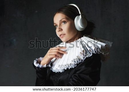 Classics are eternal, art is timeless. young woman in medieval collar and suit listens to music using modern wireless headphones, funny compilation, modernity and stylization of Renaissance paintings.