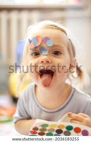 Portrait of a little lovely girl. Cute toddler sticks stickers on herself. Lovely blonde baby in a blue shirt. Royalty-Free Stock Photo #2036183255