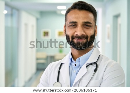Smiling bearded male Indian doctor wearing medical coat looking at camera. Headshot portrait of ethnic hispanic man medic professional, hospital physician, confident practitioner or surgeon at work. Royalty-Free Stock Photo #2036186171