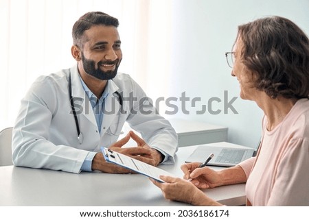 Happy Indian male doctor consulting senior old patient filling form at consultation. Friendly professional physician talking to mature woman signing medical paper during appointment visit in clinic.