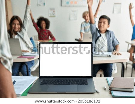 Laptop computer white blank empty mockup screen on teachers table with elementary junior children students raising hands in classroom background. Education software website technology ads concept.