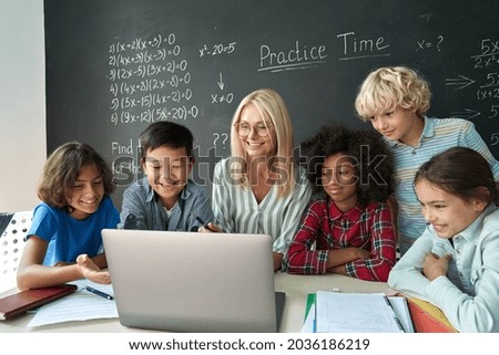 Happy diverse junior school children students gathered at teacher table looking at laptop computer using online software learning web education technology studying together at math class in classroom. Royalty-Free Stock Photo #2036186219