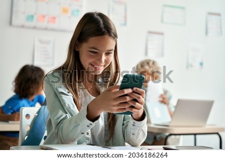 Happy latin hispanic kid girl school student using smartphone in classroom. Preteen child holding mobile cell phone having fun with apps playing games and checking social media at school during break.