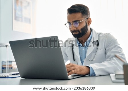 Indian male doctor medic expert wearing white coat using laptop computer at work in hospital, checking patients electronic files medical forms, having online virtual telehealth consultation. Royalty-Free Stock Photo #2036186309