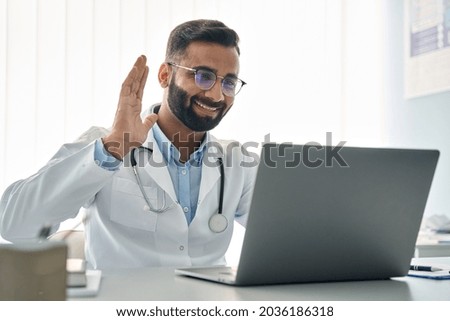Happy Indian male doctor waving hand having virtual video call online consultation on laptop computer in hospital. Tele health medicine. Telemedicine patients appointment distance remote chat in India Royalty-Free Stock Photo #2036186318
