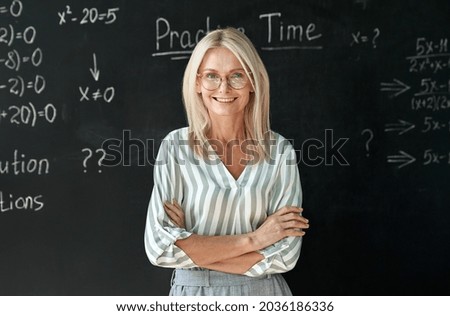 Happy smiling middle aged woman mature high school education math teacher or college professor wearing glasses standing at blackboard background. Confident female professional educator portrait.