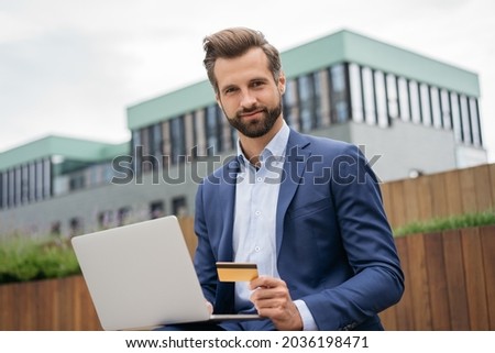 Confident businessman holding golden credit card, using laptop computer for online shopping with sales. Handsome freelancer taking payment, sitting at workplace. Successful business 	