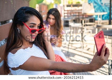 Latin girl taking a selfie for her social media. Hispanic woman with friends in the background. Young brunette woman in a summer day. Beautiful girl in a outdoor terrace. Girl with red glasses.