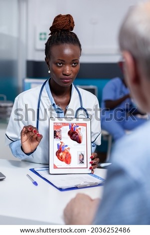 Black woman with doctor profession showing organs illustration on digital tablet display sitting at desk. African american medic explaining diagnosis to elderly sick patient in cabinet
