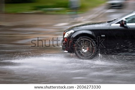 Car driving fast through big puddle at heavy rain, water splashing over the car. Car driving on asphalt road at heavy rain. Dangerous driving conditions. Dangers of aquaplaning. MOTION BLUR Royalty-Free Stock Photo #2036273330