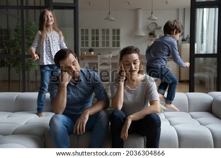 Exhausted tired couple of parents touching heads while two hyperactive noisy naughty kids jumping on couch, shouting, laughing, playing active games, having fun. Parenthood, upbringing, quarantine Royalty-Free Stock Photo #2036304866