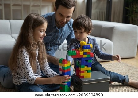 Engaged millennial dad teaching little son and daughter to build tower from construction blocks, completing bridge model from plastic bricks together, playing on heating floor at cozy home Royalty-Free Stock Photo #2036304890