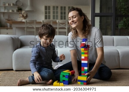 Happy best mom helping cute toddler son to build high toy tower from plastic construction blocks, playing with kid on carpet and heating floor at home, relaxing, enjoying leisure and motherhood Royalty-Free Stock Photo #2036304971
