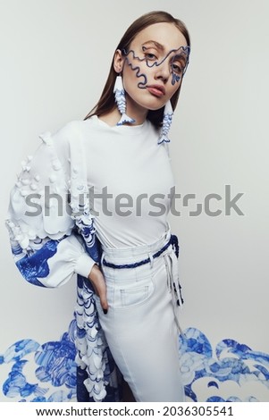 Stylish woman in wire mask and dress with blue and white waves. Drawing and ruffles on clothes. Model standing on the blue waves drawing                                        